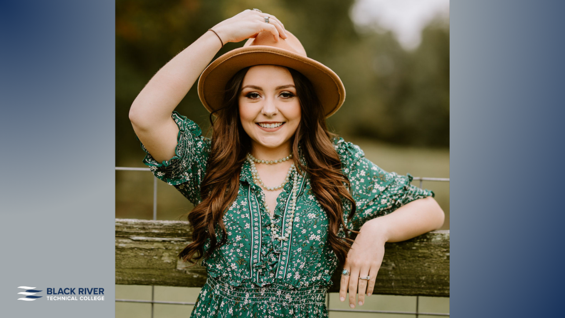 A young woman with long brown hair, wearing a green floral dress and a tan hat, smiles while leaning on a wooden fence rail outdoors, with Black River Technical College branding.